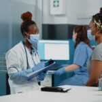 doctor-patient-with-face-masks-talking-about-disease-annual-checkup-visit-examination-medic-consulting-young-woman-cabinet-while-having-protection-against-coronavirus (1)